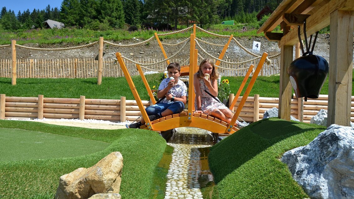 Zwei Kinder sitzen auf einer Holzbrücke und genießen Eiscreme. Im Hintergrund sieht man eine grüne Landschaft mit Bäumen und Spielbereichen. | © Mountain Adventure Golf