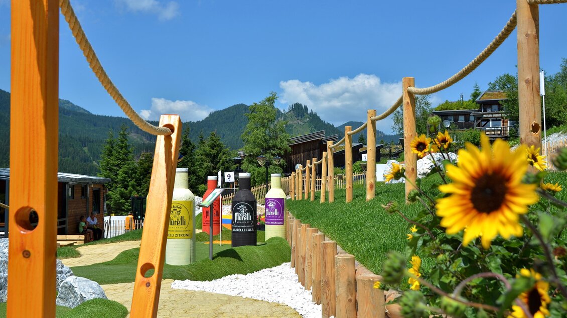 Ein Weg mit Holzgeländern führt durch eine grüne Landschaft. Im Hintergrund sind Getränke-Displays und Sonnenblumen zu sehen, während die Berge und der klare Himmel für eine schöne Kulisse sorgen. | © Mountain Adventure Golf