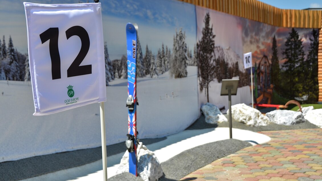 Ein Skiareal mit einem Hinweisschild Nummer 12 und einem Skiset. Im Hintergrund sind verschneite Bäume und eine winterliche Landschaft zu sehen. | © Mountain Adventure Golf