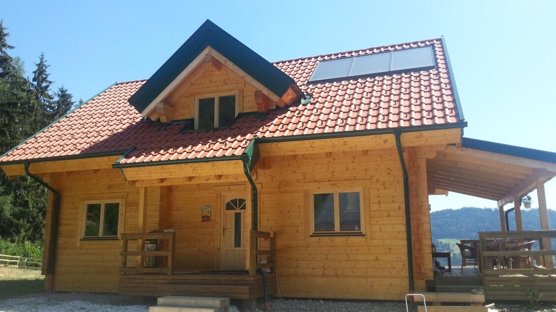 Ein modernes Holzhaus mit rotem Ziegeldach und großer Veranda. Es steht in einer hügeligen Landschaft mit Bäumen im Hintergrund. | © Murtal Alpaka
