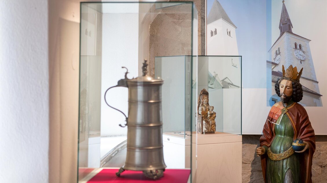 Ein antiker Krug steht in einer Vitrine, umgeben von einer hellen Ausstellungsumgebung. Zu sehen ist außerdem eine Statue einer Königin im Hintergrund. | © Stadtgemeinde Kapfenberg