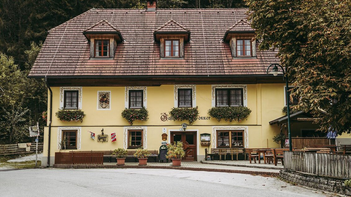 Ein charmantes, gelbes Haus mit einem roten Ziegeldach und Fenstern, die mit Blumen dekoriert sind. Vor dem Haus stehen Tische und Stühle für Gäste.