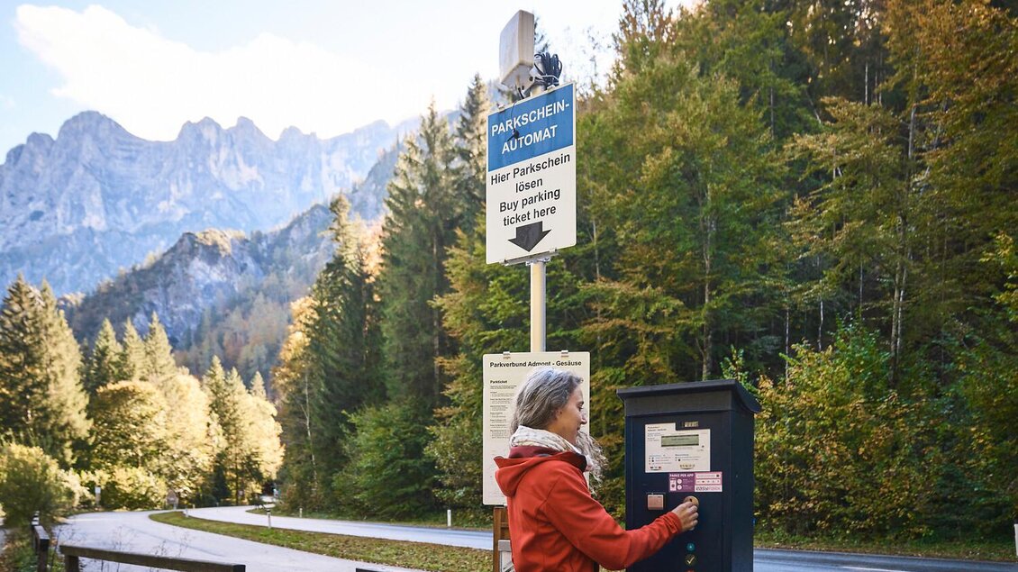 Eine Frau steht an einem Parkautomaten in den Bergen. Im Hintergrund sind hohe Berge und bunte Herbstbäume zu sehen.