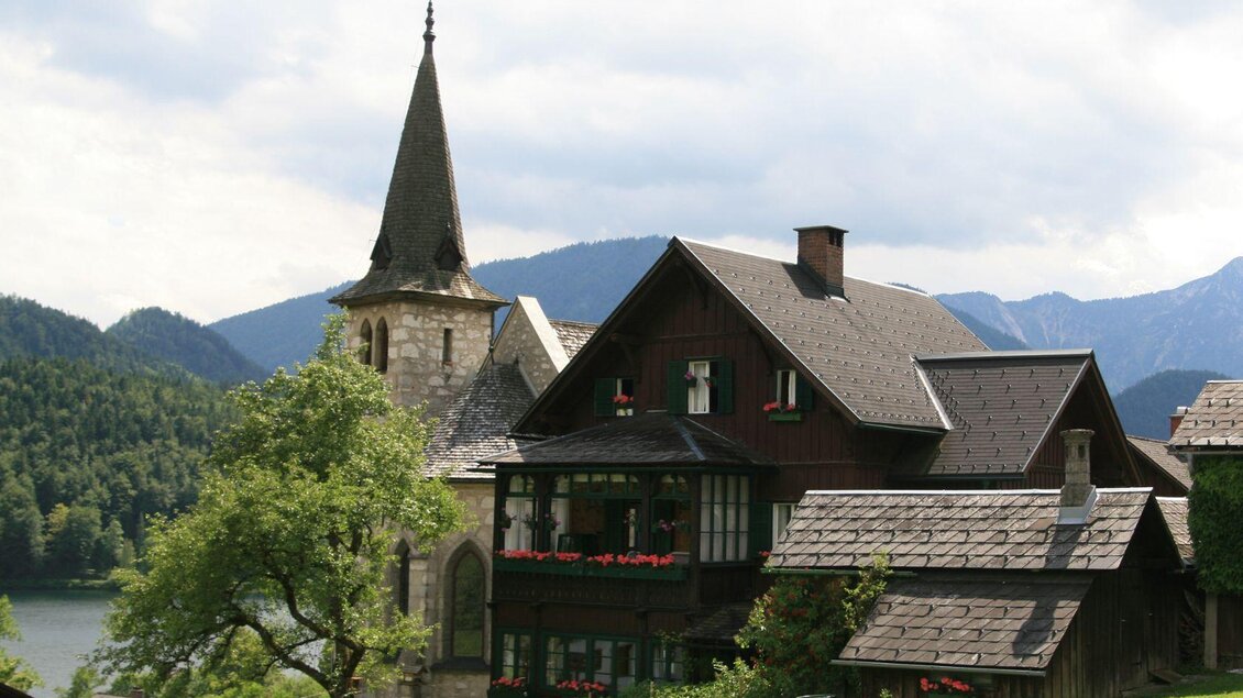 Ein malerisches Dorfhaus mit einem hohen Kirchturm und blühenden Fenstern. Im Hintergrund sind sanfte Berge und ein ruhiger See zu sehen. | © Viola Lechner