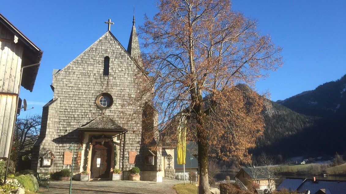 Eine kleine Kirche aus Holz mit einem spitzen Dach und einem Baum im Vordergrund. Im Hintergrund sind Berge und ein klarer blauer Himmel sichtbar. | © Bettina Scheck
