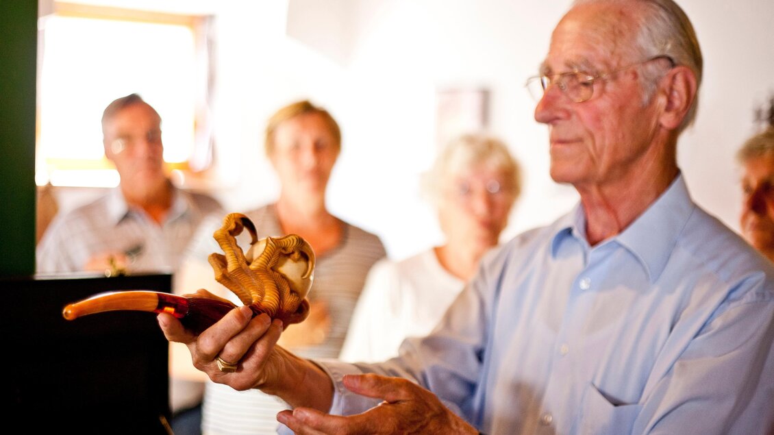 Ein älterer Mann hält ein kunstvolles Holzobjekt in der Hand. Im Hintergrund sind mehrere Personen zu sehen, die aufmerksam schauen. | © Holzwelt Murau