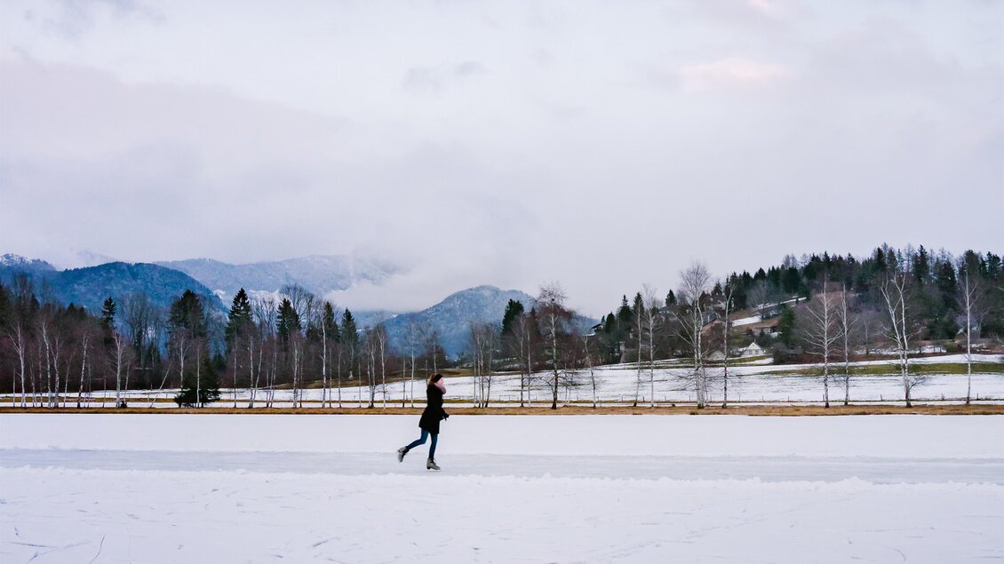 Eine Eisläuferin bewegt sich über eine gefrorene Fläche im Winter. Im Hintergrund sind schneebedeckte Berge und Bäume zu sehen. | © Christoph Lukas