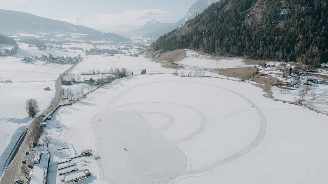 Eine weite verschneite Landschaft mit einem eisbedeckten See. Im Hintergrund sind Berge und ein klarer Himmel zu sehen.