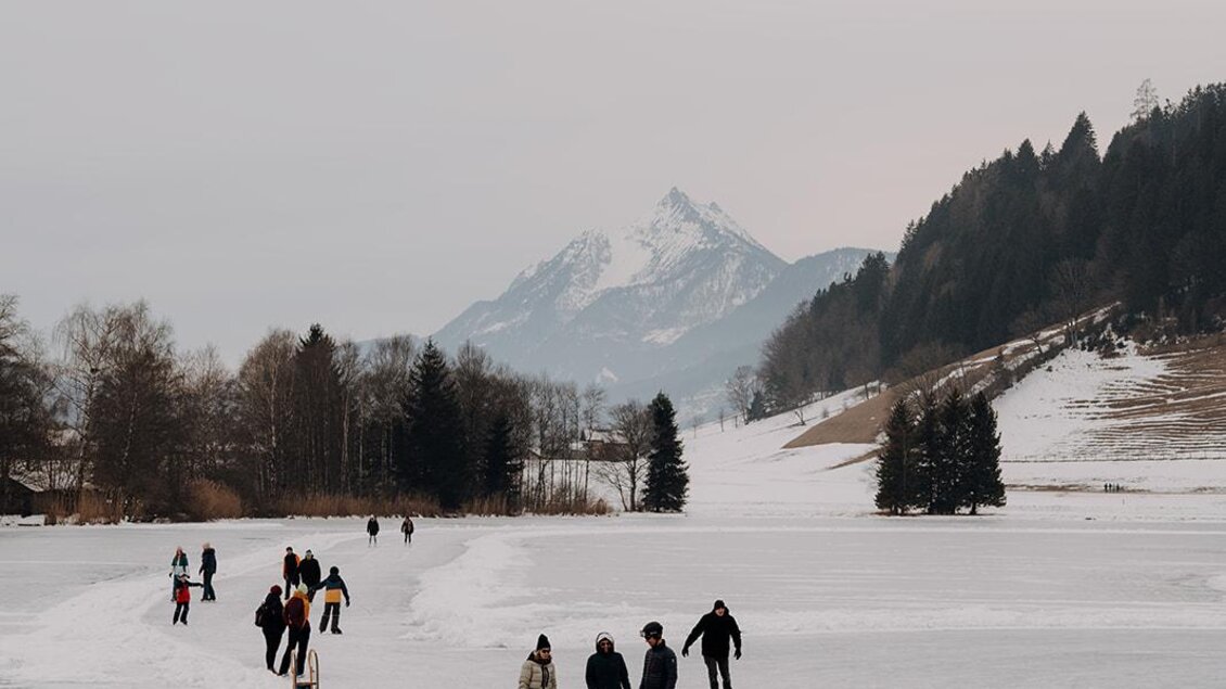 Menschen laufen auf einer gefrorenen Fläche im Schnee. Im Hintergrund sind Berge und eine bewölkte Himmel zu sehen.