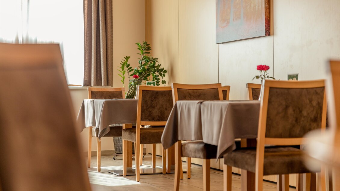 Ein gemütliches Restaurant mit Holzstühlen und Tischen, die mit grauen Tischdecken bedeckt sind. Im Hintergrund sind Fenster und eine Pflanzenanordnung zu sehen. | © Restaurant Leitner