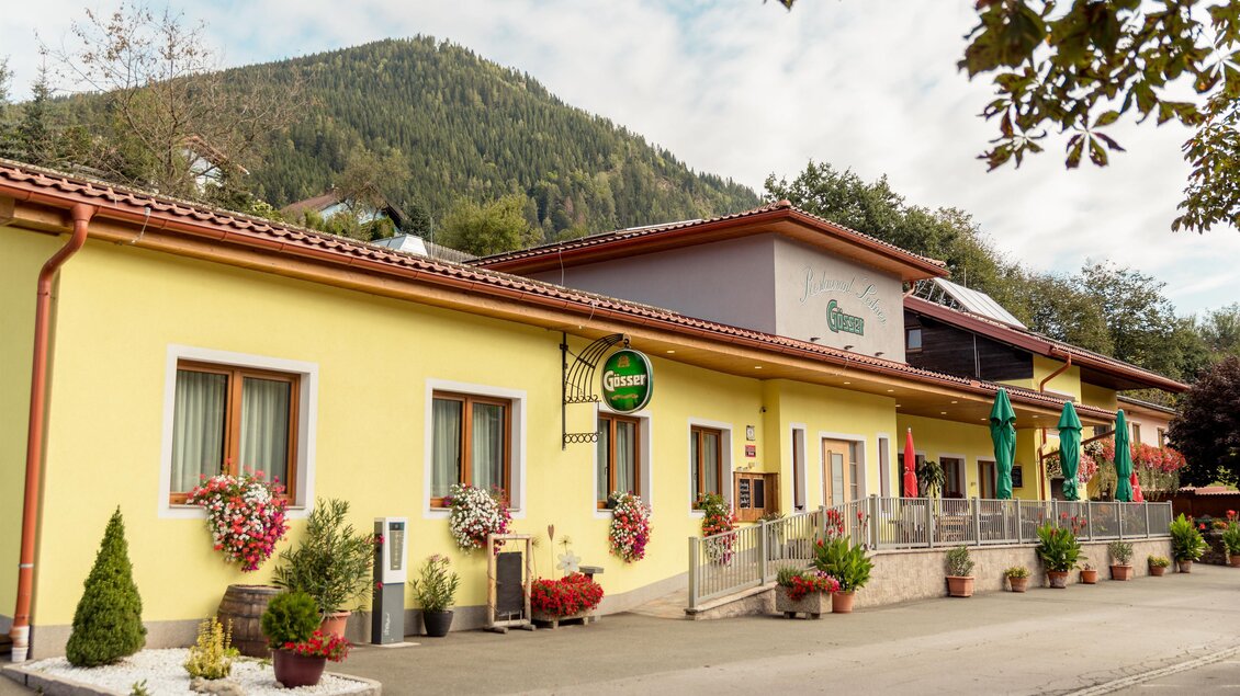 Ein charmantes Haus im Landhausstil mit gelber Fassade und blühenden Blumenkästen. Im Hintergrund sind sanfte Hügel und Bäume zu sehen. | © Restaurant Leitner