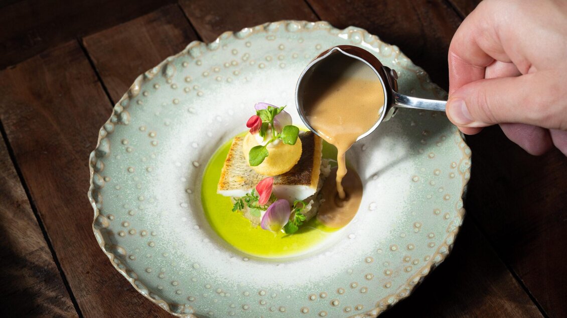 Gebratenes Fischfilet mit Kräutern und Blüten auf grünem Saucenspiegel, während eine Hand braune Sauce darüber gießt im Restaurant Wörgötter in Ligist.  | © Die Abbilderei