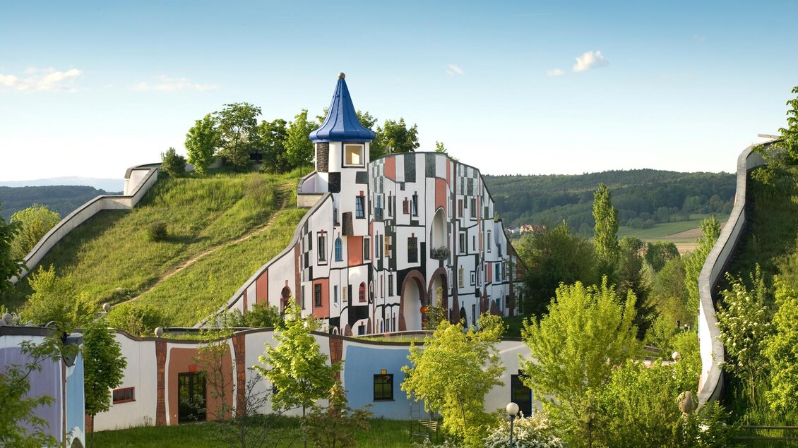 Ein buntes Gebäude auf einem Hügel mit einem blauen Turm. Die Landschaft ist grün und von Bäumen umgeben. | © Rogner Bad Blumau/Hundertwasser Architekturprojekt