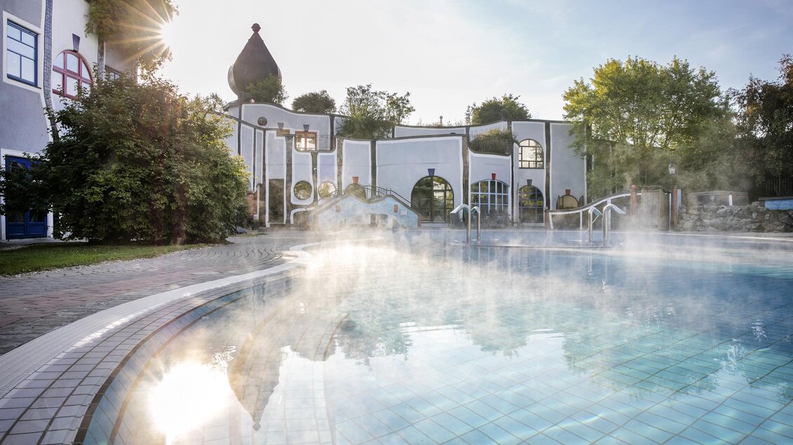 Ein ruhiger Wellnessbereich mit einem dampfenden Außenpool. Die Sonne strahlt hinter der Architektur und umgeben von grünen Bäumen. | © Rogner Bad Blumau/Hundertwasser Architekturprojekt