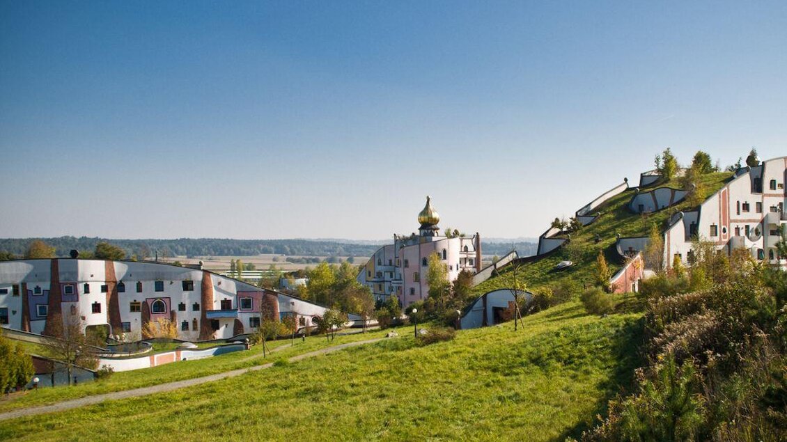 Eine malerische Landschaft mit Hügeln und bunten Gebäuden. Im Hintergrund erstreckt sich der Himmel über einer weiten Ebene. | © Rogner Bad Blumau/Hundertwasser Architekturprojekt