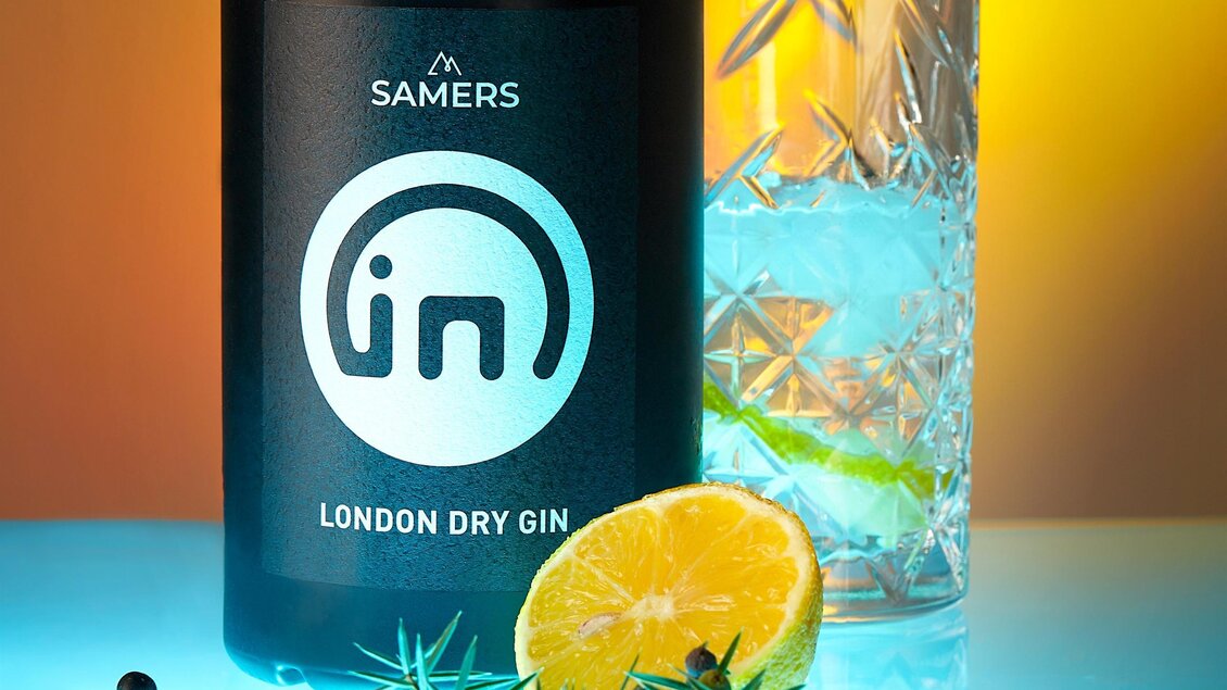 Ein Glas mit Eis und einer Flasche London Dry Gin stehen auf einem reflektierenden Tisch. Frische Kräuter und eine Zitronenscheibe ergänzen das Bild. | © photoINstyle