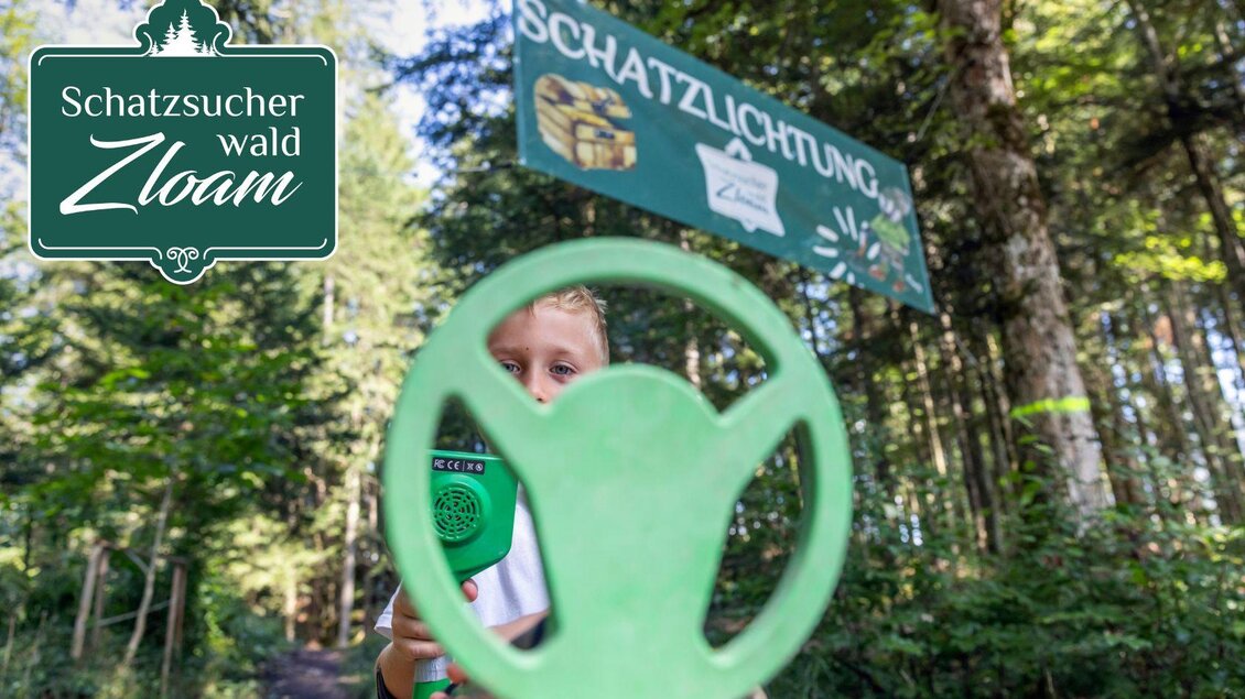 Ein Kind hält eine grüne Schatzsuche-Sonde vor einem Waldweg. Im Hintergrund ist ein Schild mit der Aufschrift "Schatzlichtung" zu sehen. | © www.zloam.at