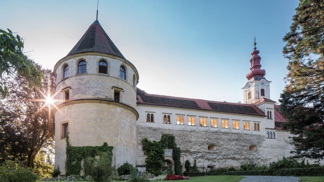 Eine historische Burg mit einer markanten Turmstruktur und grünen Gärten. Die Sonne scheint sanft hinter dem Gebäude hervor. | © Leonhard Hirzensauer