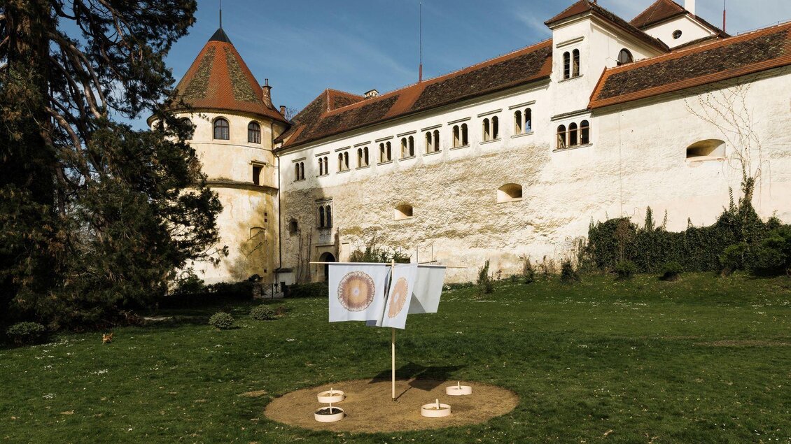 Eine historische Burg mit einem grünen Garten. Im Vordergrund hängen Bilder an einer Leine. | © Lupi Spuma