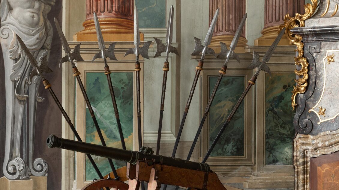 Eine Kanone auf Rädern steht in einem historisch eingerichteten Raum. Im Hintergrund sind Säulen und klassische Statuen zu sehen. | © LippZahnschirm