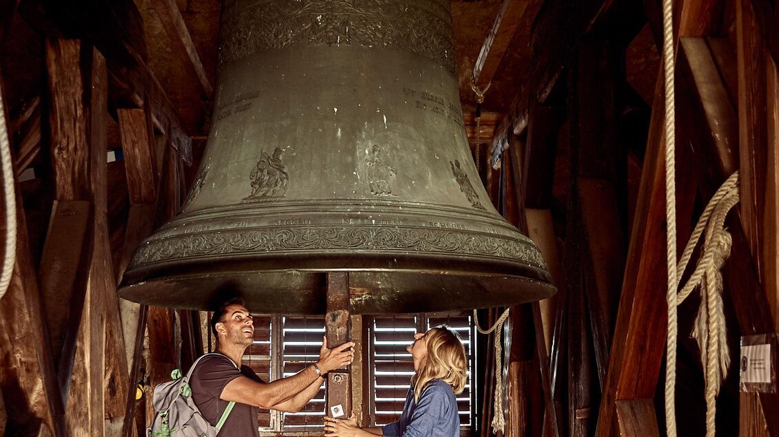 Eine große Glocke hängt in einem Holzgerüst. Zwei Personen stehen darunter und betrachten sie. | © Stephan Friesinger