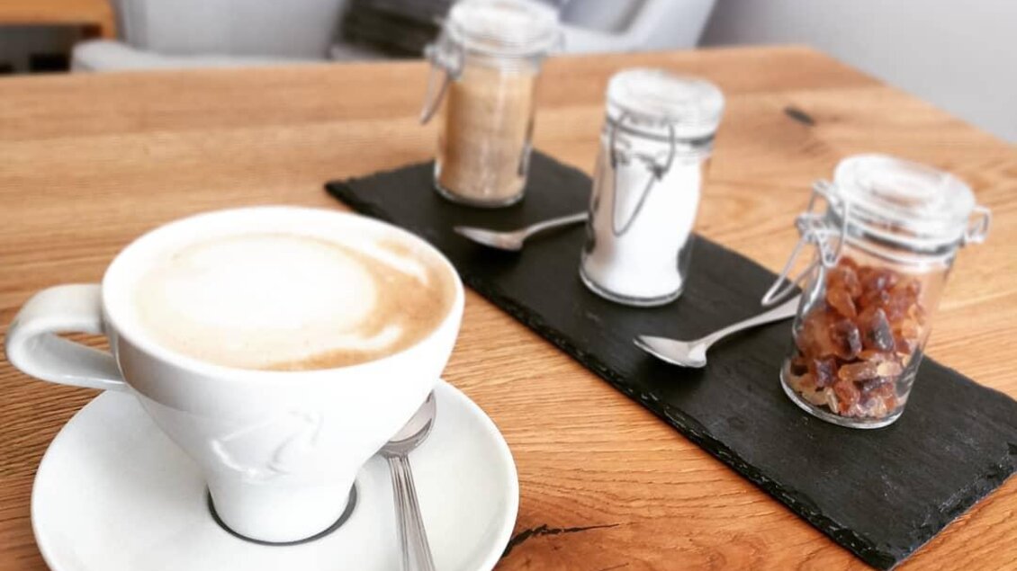 Eine Tasse Kaffee mit Milchschaum steht auf einem Holztisch. Neben der Tasse befinden sich drei Gläser mit Zucker und Süßstoff.