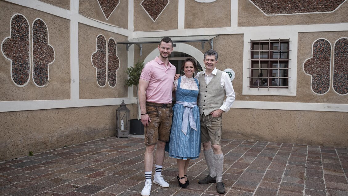 Drei Personen in traditioneller bayerischer Tracht stehen vor einer historischen Wand. Sie lächeln und posieren freundlich für das Foto. | © Schloss Farrach