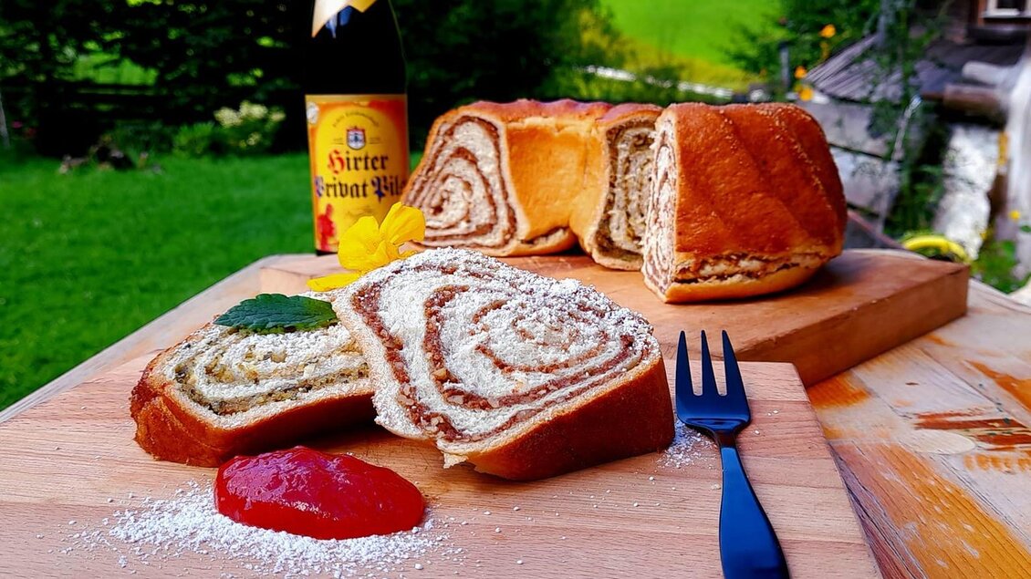 Ein köstlicher Zopf mit Marmelade und Puderzucker auf einem Holzbrett. Im Hintergrund sieht man eine grüne Wiese und einen Bierflasche. | © Brudermann