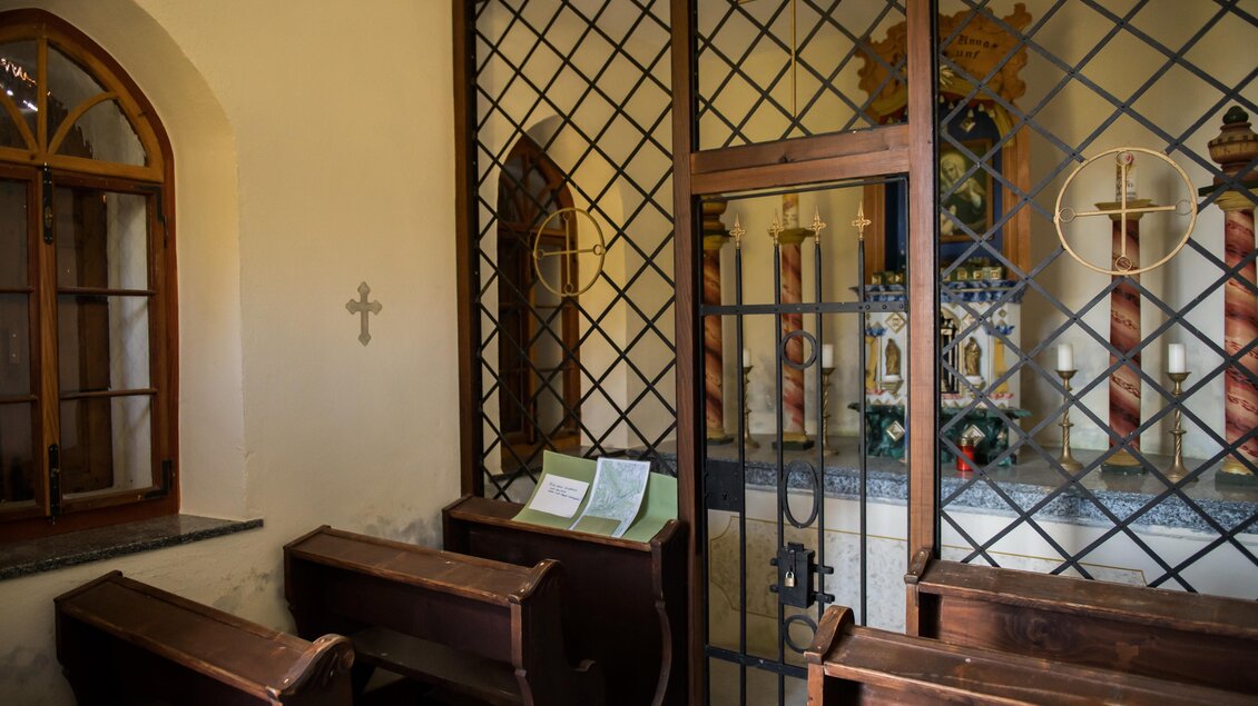 Ein kleiner, ruhiger Innenraum einer Kirche mit Holzbänken und einem Fenster. Im Hintergrund befindet sich ein Altar, der von Gitterstäben umgeben ist. | © Anita Fössl