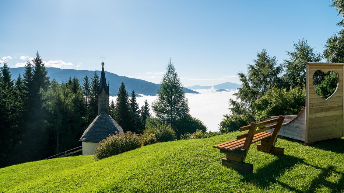 Eine idyllische Landschaft mit einer grünen Wiese, Bäumen und einem Blick auf Nebel im Tal. Im Hintergrund sind Berge und eine kleine Kapelle sichtbar. | © Anita Fössl