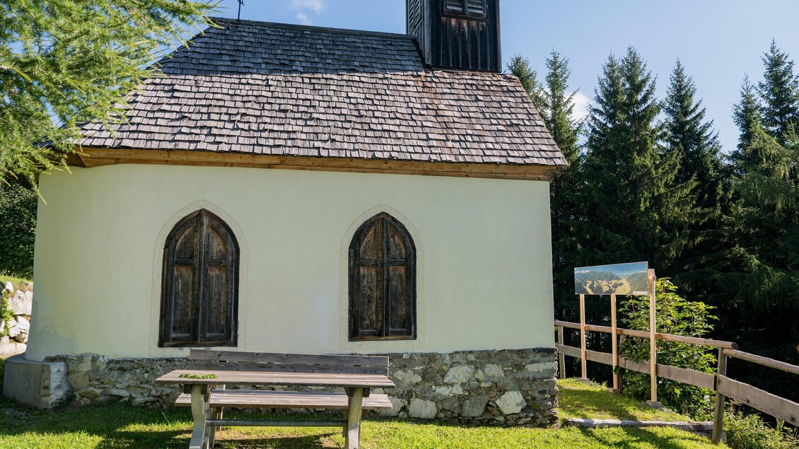 Eine kleine, charmante Kapelle mit Holzfenstern steht inmitten von Bäumen. Das grüne Gras und der klare Himmel schaffen eine friedliche Atmosphäre. | © Anita Fössl