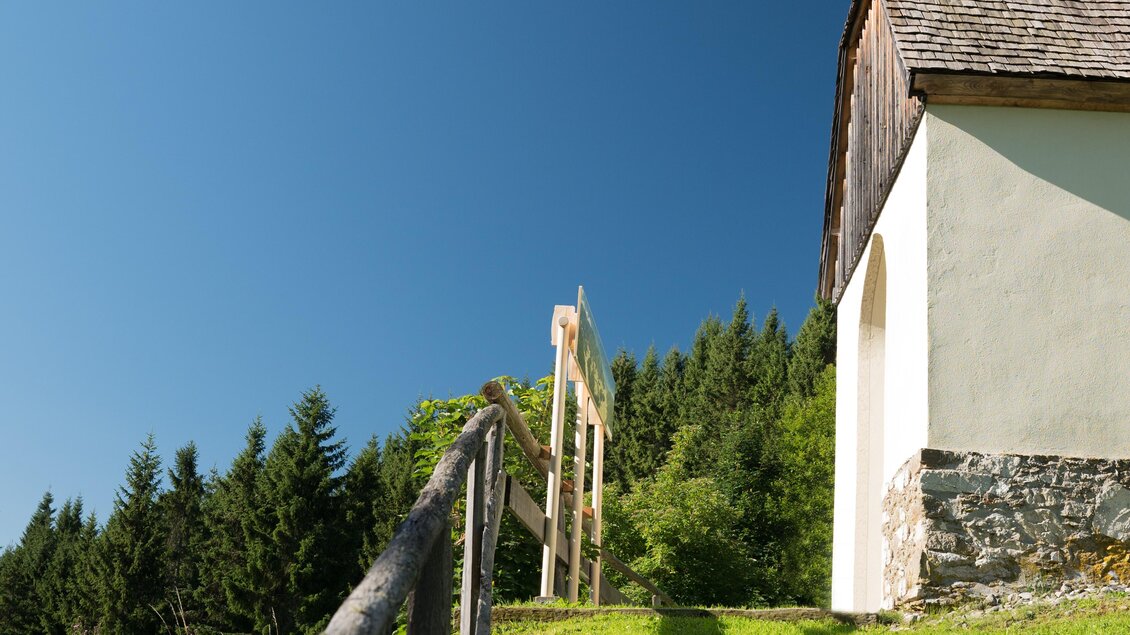 Eine kleine Kirche steht auf einem Hügel mit grünem Gras und einem klaren blauen Himmel. Der Weg zur Kirche wird von einem Geländer gesäumt. | © Anita Fössl