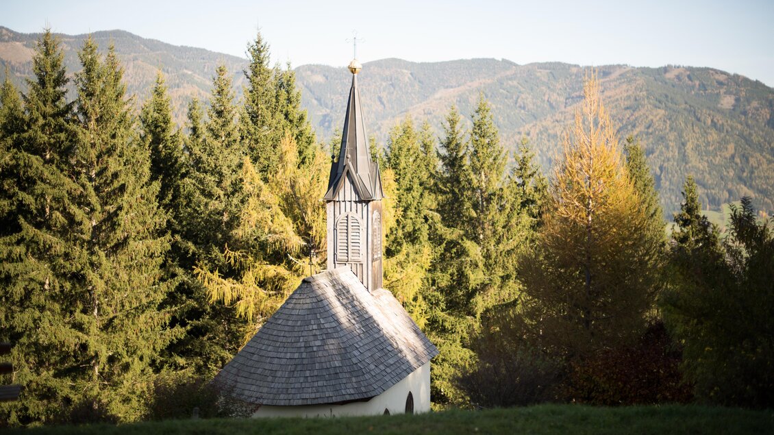 Eine kleine Kapelle mit einem spitzen Turm steht zwischen hohen Bäumen. Im Hintergrund sind sanfte Hügel und Berge zu sehen. | © Anita Fössl