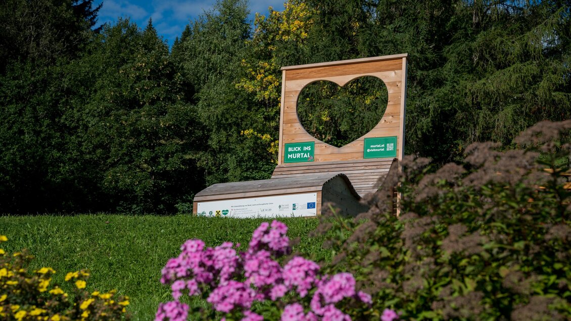 Ein großes Herz aus Holz steht in einer grünen Landschaft. Vor dem Herz blühen bunte Blumen. | © Anita Fössl