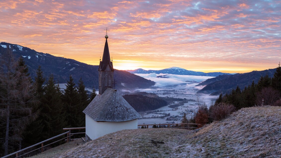 Eine malerische Berglandschaft mit einer kleinen Kirche im Vordergrund. Der Himmel ist in warmen Farben bei Sonnenaufgang gefärbt, und Nebel liegt in den Tälern. | © Markus Beren