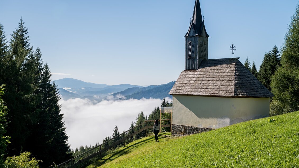 Eine kleine Kirche auf einer grünen Wiese mit Bäumen im Hintergrund. Über der Landschaft liegt ein Nebelteppich, während die Berge in der Ferne sichtbar sind. | © Anita Fössl
