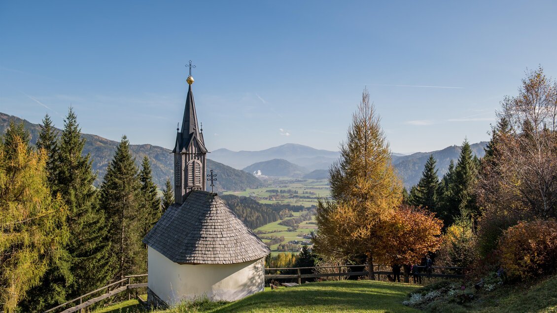 Eine kleine Kapelle steht auf einer grünen Wiese, umgeben von Bäumen und Bergen. Der Himmel ist klar und die Landschaft strahlt in warmen Herbstfarben. | © Anita Fössl