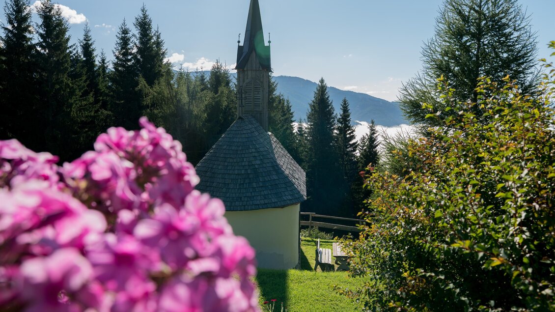 Eine kleine Kirche umgeben von Bäumen und bunten Blumen. Im Hintergrund sind Berge und ein klarer Himmel zu sehen. | © Anita Fössl