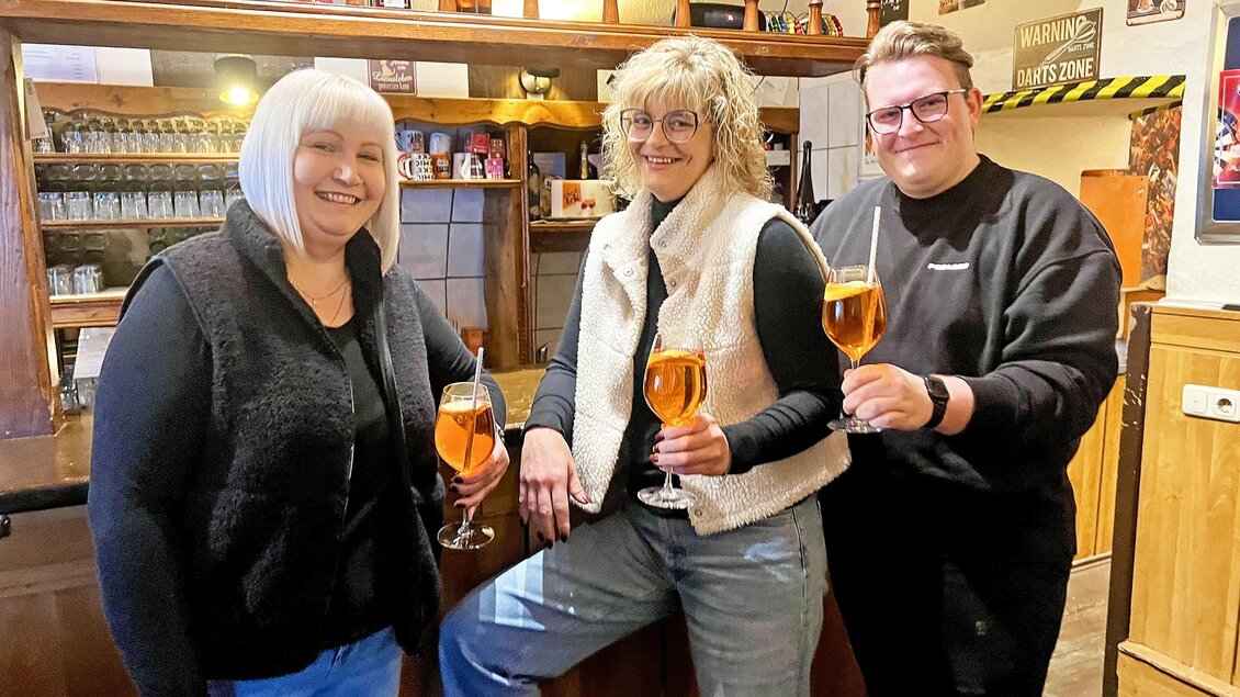 Drei Personen stehen an einer Bar und halten Gläser mit Getränken in der Hand. Sie lächeln freundlich in die Kamera. | © Stadtmarketing Judenburg