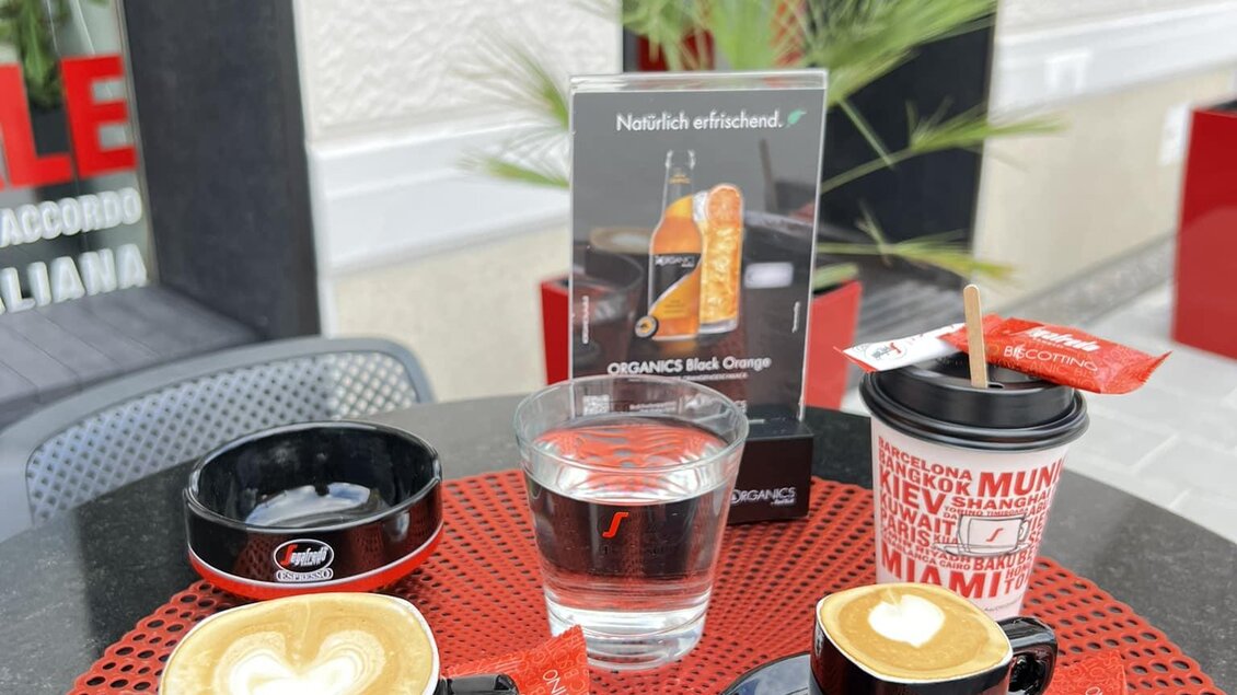 Ein Tisch mit zwei Tassen Kaffee, einem Wasserglas und einem Getränkebecher. Im Hintergrund ist eine Pflanze und ein Schild sichtbar. | © Andi Bäuchl