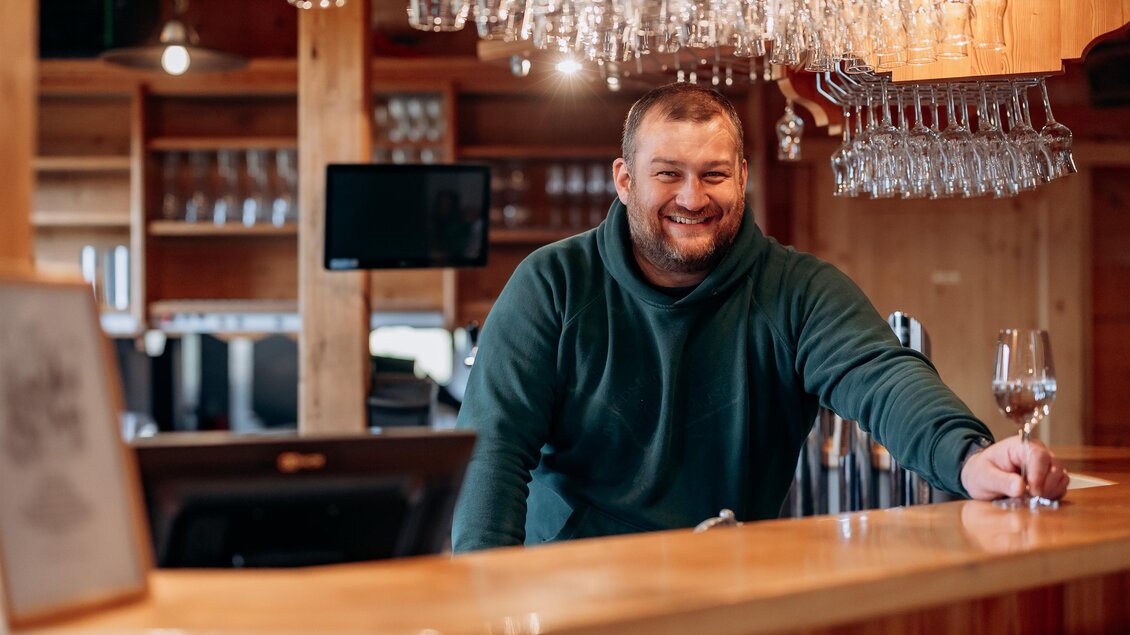 Ein lächelnder Barkeeper steht hinter der Theke und hält ein Weinglas. Im Hintergrund hängen Gläser an der Decke, und die Atmosphäre ist einladend. | © Stift Admont/Christoph Draxl