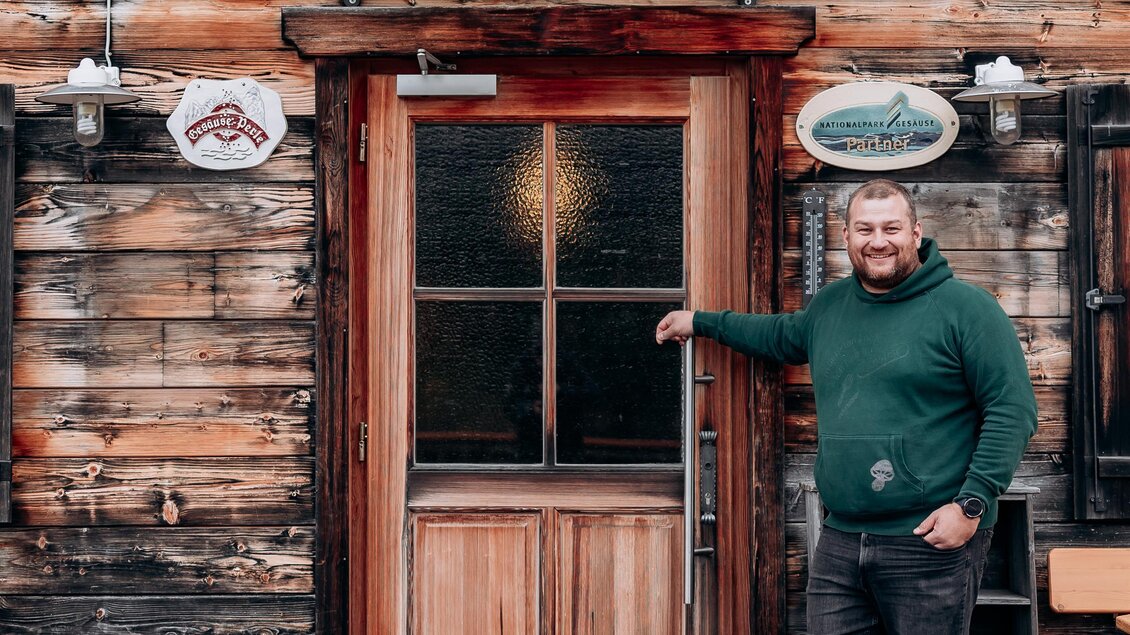Ein Mann steht vor dem Eingang zur SportAlm, einem rustikalen Holzgebäude. Das Fenster ist gut sichtbar und das Schild über der Tür zeigt den Namen des Lokals. | © Stift Admont/Christoph Draxl
