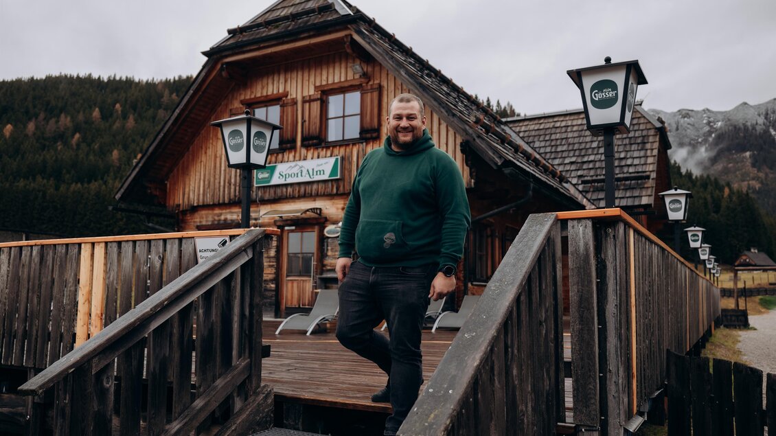 Ein Mann steht auf der Treppe vor einem traditionellen Holzhaus in den Bergen. Der Himmel ist bewölkt und die Umgebung ist naturnah. | © Stift Admont/ Christoph Draxl