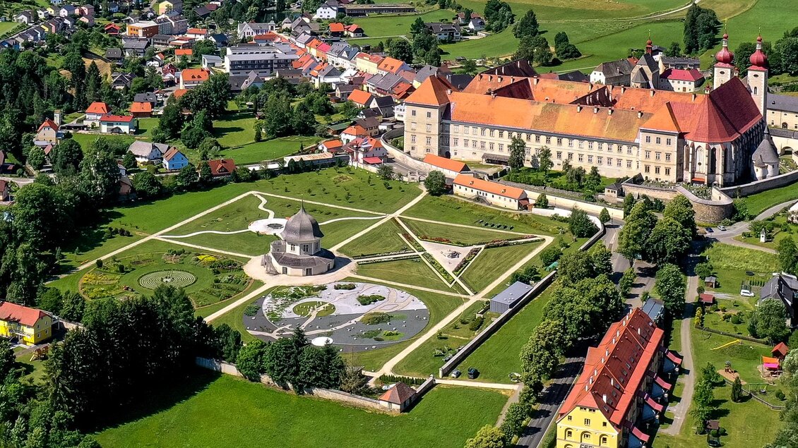 Eine Luftaufnahme eines historischen Schlosses mit gepflegten Gärten und einem großen Park. Umgeben von einem hübschen Dorf mit bunten Häusern im Hintergrund. | © Holzwelt Murau