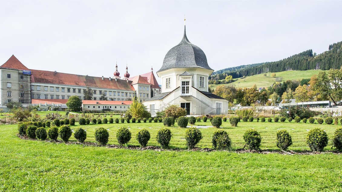Ein wunderschönes Gebäude mit einem hohen Dach steht in einem gepflegten Garten. Im Hintergrund sind sanfte Hügel und eine historische Anlage zu sehen. | © TVB Murau