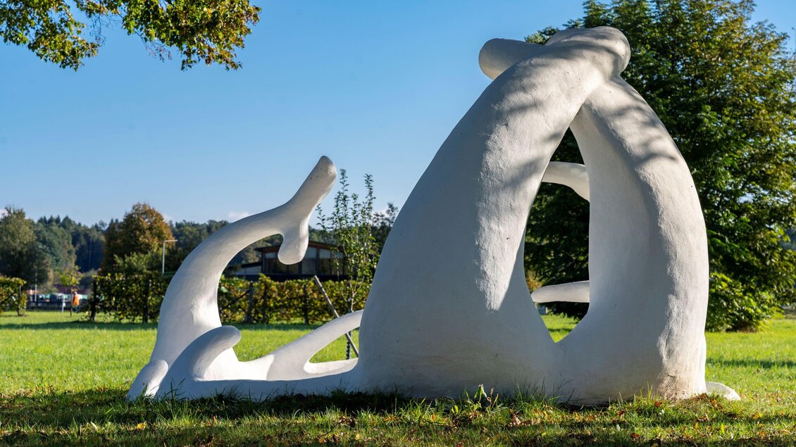 Eine moderne Skulptur aus weißem Material steht in einer grünen Wiese. Im Hintergrund sind Bäume und ein klarer blauer Himmel zu sehen. | © Kurkommission Bad Blumau