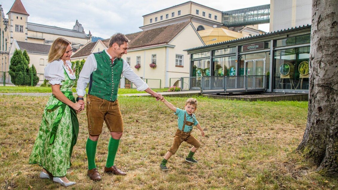Eine Familie in traditioneller Tracht steht im Freien. Sie hält miteinander Händchen und lächelt, während ein Kind spielt. | © TVB Erzberg-Leoben