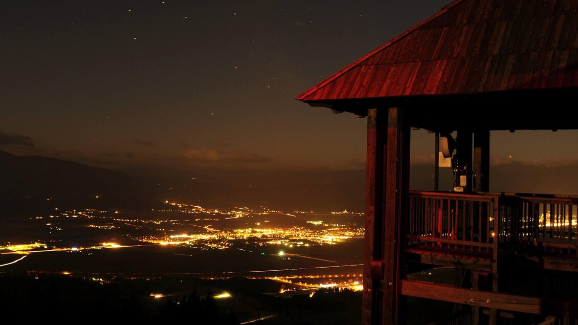 Eine Aussicht von einem Hügel bei Nacht, mit einem Holzturm und Stadtlichtern im Hintergrund. Der Himmel ist dunkel mit vereinzelten Sternen. | © Peter Eberhard