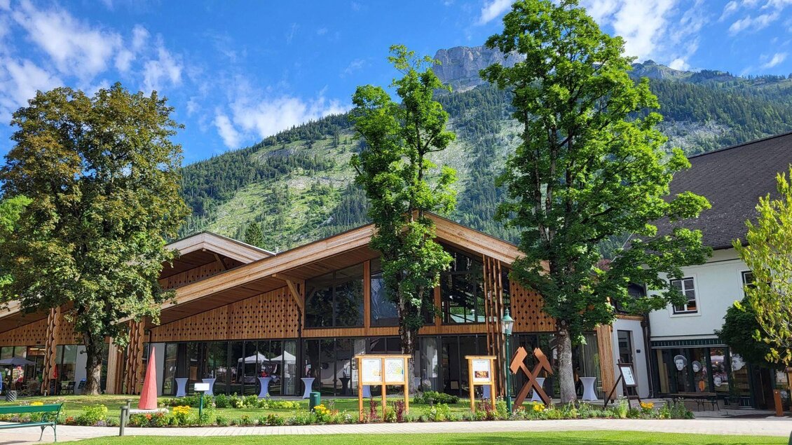 Der moderne Veranstaltungssaal im Herzen von Altaussee aus viel Holz und Glas. Im Vordergrund ist der grüne Kurpark der Gemeinde am Altausseer See zu sehen, der von einer Skulptur mit dem Bergmannszeichen aus Cortenstahl in seiner charakteristischen rostbraunen Farbe geschmückt wird, welche am rechten Bildrand zu sehen ist. | © veranstaltungssaal-altaussee-sommer-foto-petra-kirchschlager.