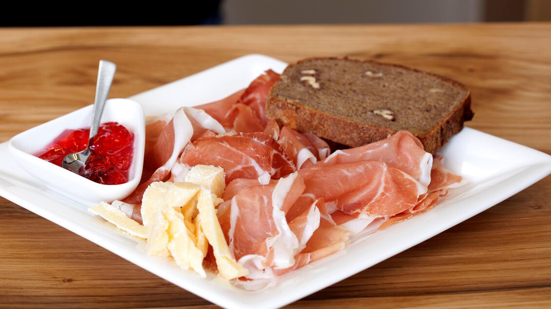 Ein Teller mit aufgeschnittenem Schinken, ein Stück Brot und einem kleinen Schälchen Marmelade. Die Anrichtung ist übersichtlich und ansprechend. | © Vulcano Schinkenmanufaktur